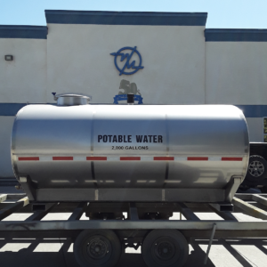 Silver portable water tank on a trailer labeled 'POTABLE WATER 2,000 GALLONS' in a parking lot in front of a building.