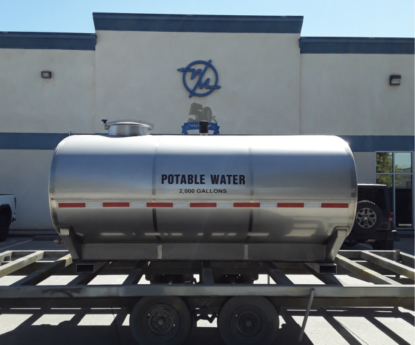 Silver portable water tank on a trailer labeled 'POTABLE WATER 2,000 GALLONS' in a parking lot in front of a building.